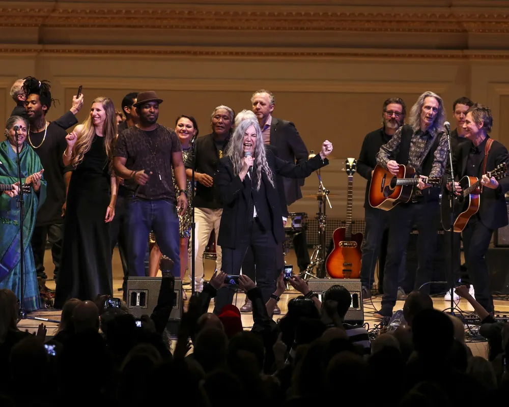 A diverse group of musicians performing together on stage at a concert hall, holding guitars and other instruments, with an audience visible in the foreground watching the performance.