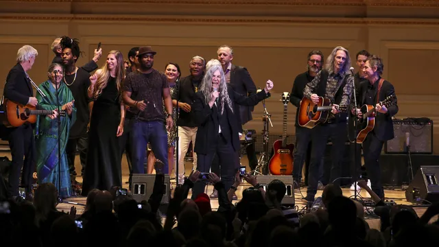 A diverse group of musicians performing together on stage at a concert hall, holding guitars and other instruments, with an audience visible in the foreground watching the performance.