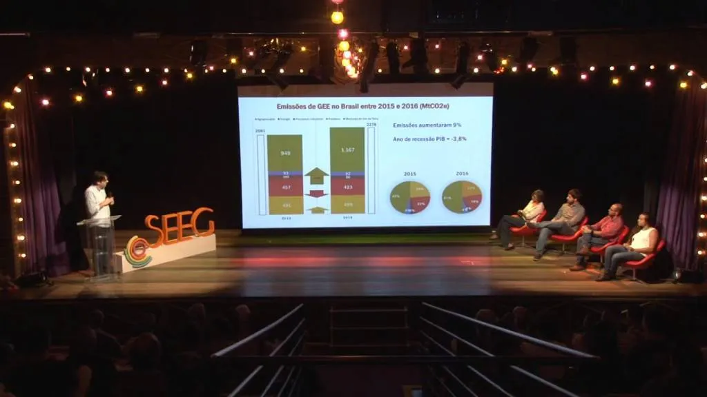 A presentation on a stage showing greenhouse gas emissions data for Brazil (2015-2016). Speaker at podium on left, panel of four seated on right. Stage decorated with "SEEG" signage and string lights.