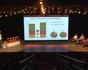 A presentation on stage showing data visualization with charts and graphs on a large screen. Speakers and audience members are seated on stage under warm string lights in what appears to be a theater or conference venue.