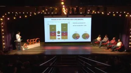 A presentation on stage showing data visualization with charts and graphs on a large screen. Speakers and audience members are seated on stage under warm string lights in what appears to be a theater or conference venue.
