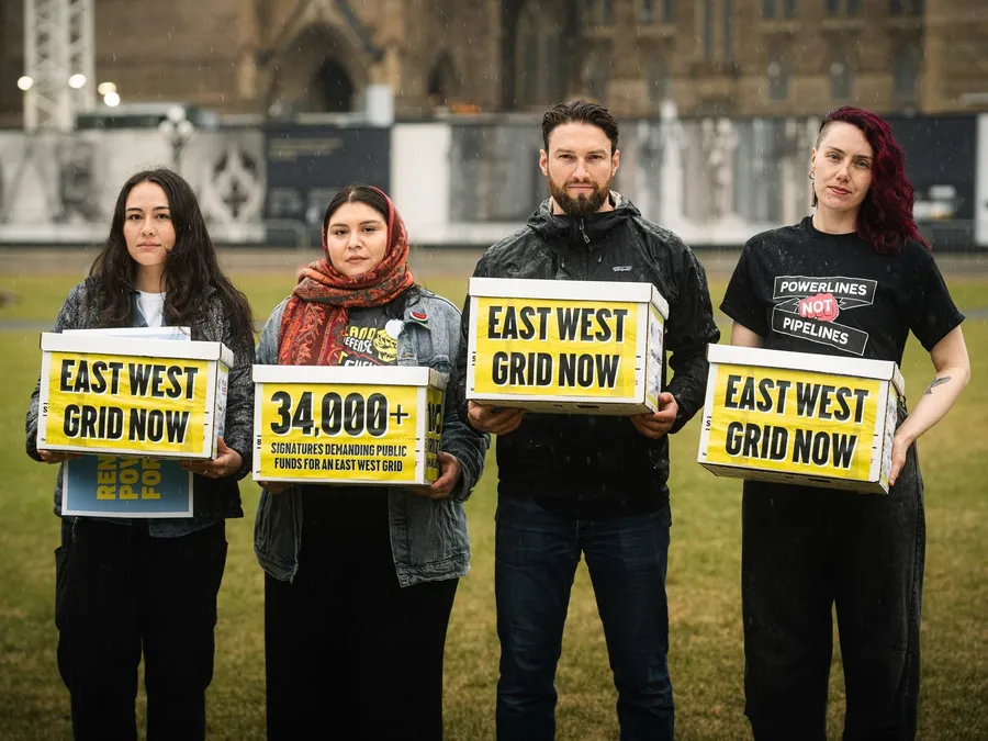 Four activists stand in front of Parliament holding "EAST WEST GRID NOW" signs and a "34,000+ signatures" petition, advocating for public investment in an East West electricity grid infrastructure project.