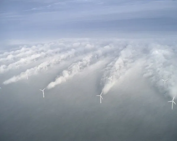 Aerial view of an offshore wind farm emerging through thick clouds or fog. White wind turbines stand in rows above a gray cloudy layer, creating a dramatic atmospheric scene over what appears to be open water.