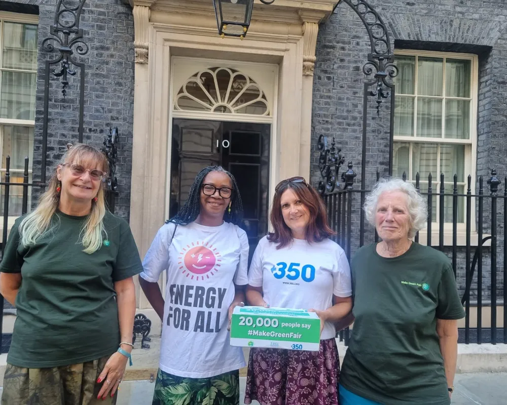Four women stand outside 10 Downing Street holding climate action signs. They wear t-shirts with environmental messages including "Energy for All" and "350". They're advocating for green energy with a petition sign showing 20,000 supporters.