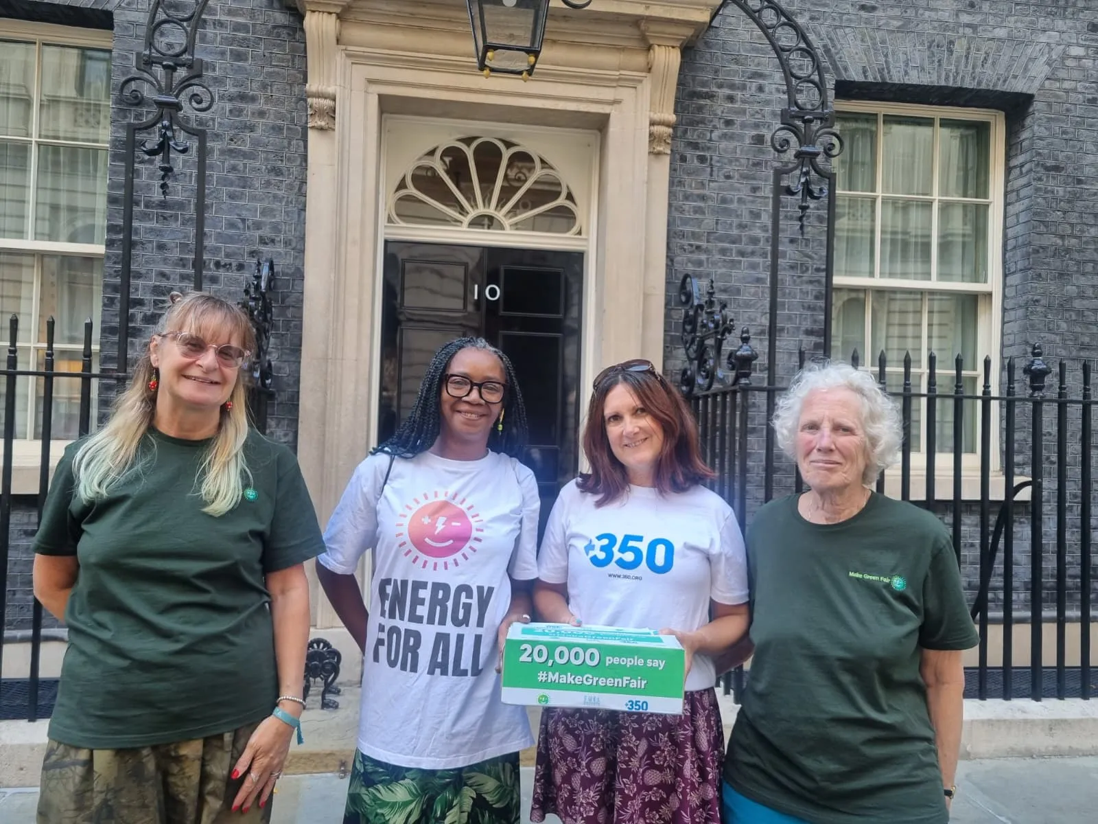 Four women stand outside 10 Downing Street holding climate action signs. They wear t-shirts with environmental messages including "Energy for All" and "350". They're advocating for green energy with a petition sign showing 20,000 supporters.