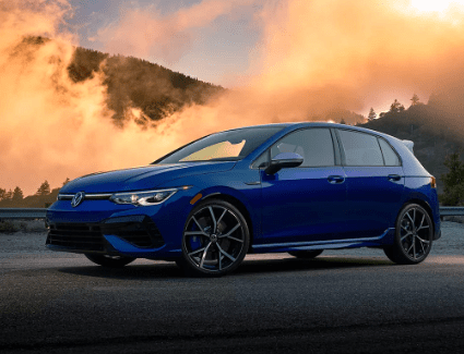 New 2023 Volkswagen Golf R exterior image Front side view of a blue 2023 Volkswagen Golf R parked on a road against a misty mountain backdrop.