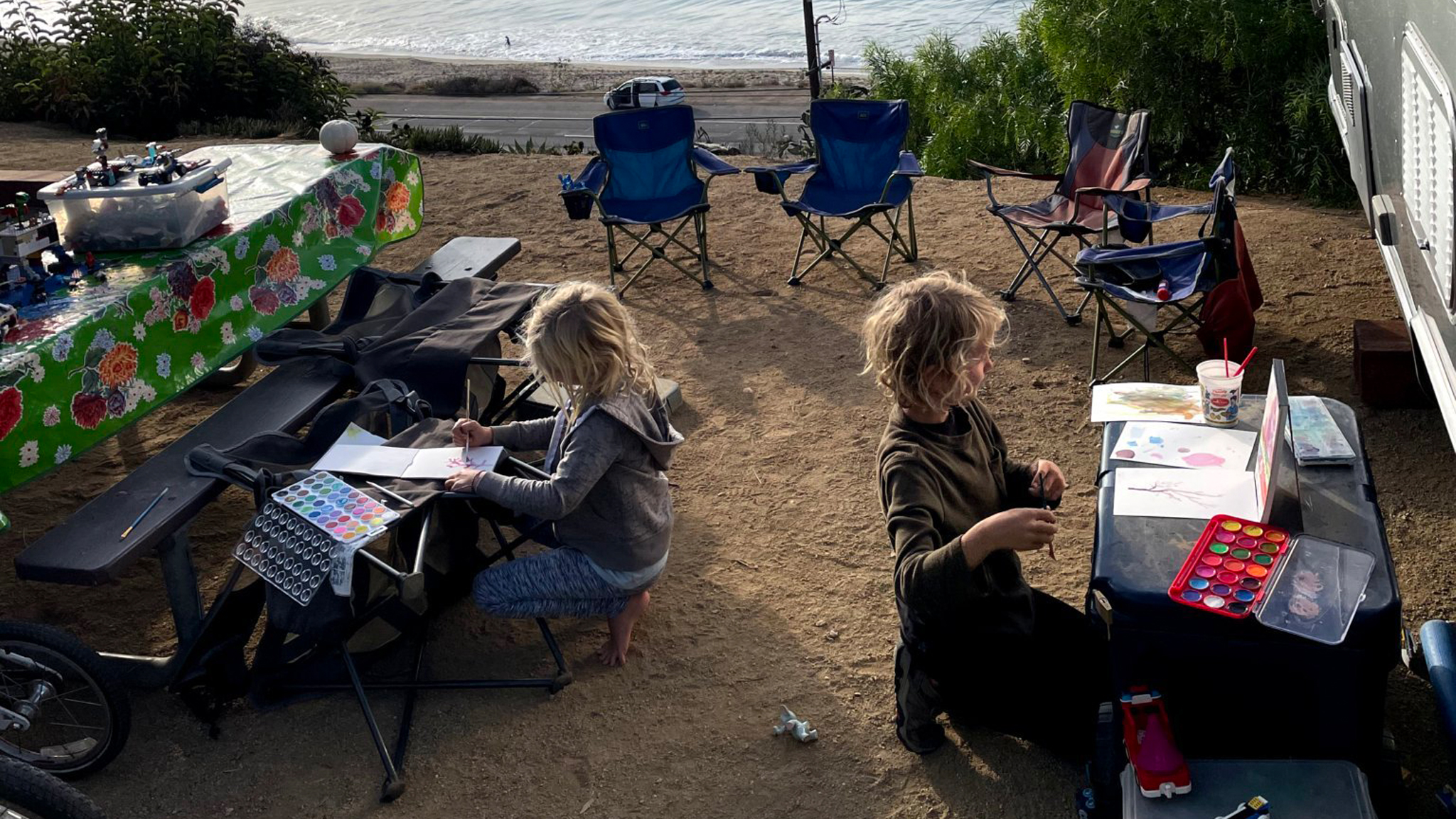 Two kids sitting outside of their Airstream travel trailer at their own tables while they paint next to the water.|Three kids playing in the ocean.|Four kids sitting on the edge of a boat with lifejackets on looking out at the water.|A young girl in a blue jacket carrying a bucket of things she found as she walks along a beach.|A young kid sitting at a picnic table writing about nature|Two kids sitting at a table outside of their Airstream travel trailer while doing crafts.|A young kid writes in a notebook about what they found in nature.|A father and two kids sitting outside of their Airstream travel trailer while they learn and write about their adventures.|Two kids sitting in the galley of their Airstream travel trailer while they learn on the road.|Two kids sitting outside of their Airstream travel trailer at their own tables while they paint next to the water.