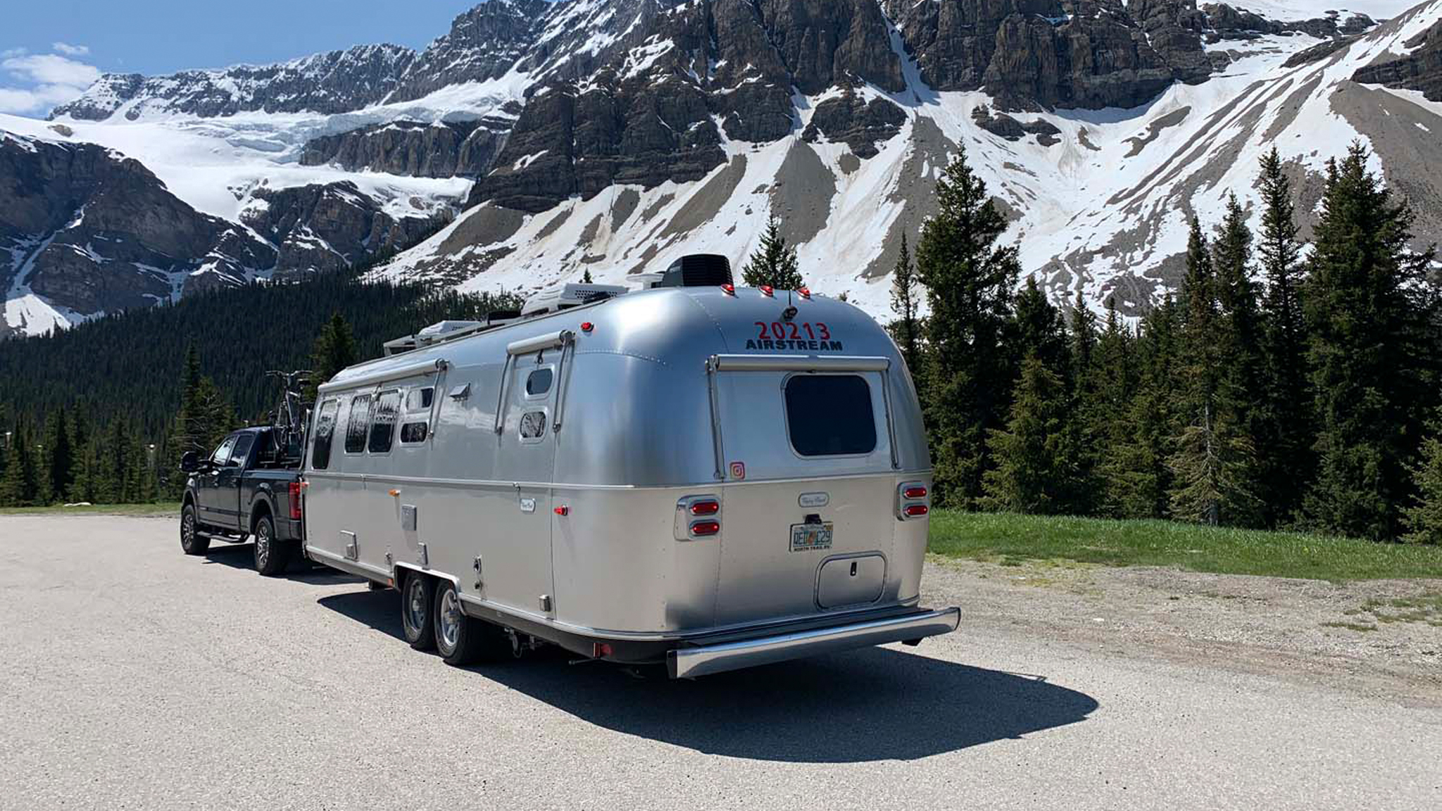 Airstream camper being towed by a truck through snowy mountains.|Airstream camper being towed by a truck through snowy mountains.|Airstream Flying Cloud Office camper boondocking.|A man sitting in the Airstream Flying Cloud Office writing music.|A space in the Flying Cloud Office camper made as a spot for a keyboard.|A man working from anywhere in his Airstream Flying Cloud office camper.|Scott and Kim posing for an image next to a lake and mountains.|Airstream Flying Cloud camper parked at a campground with trees surrounding the trailer.