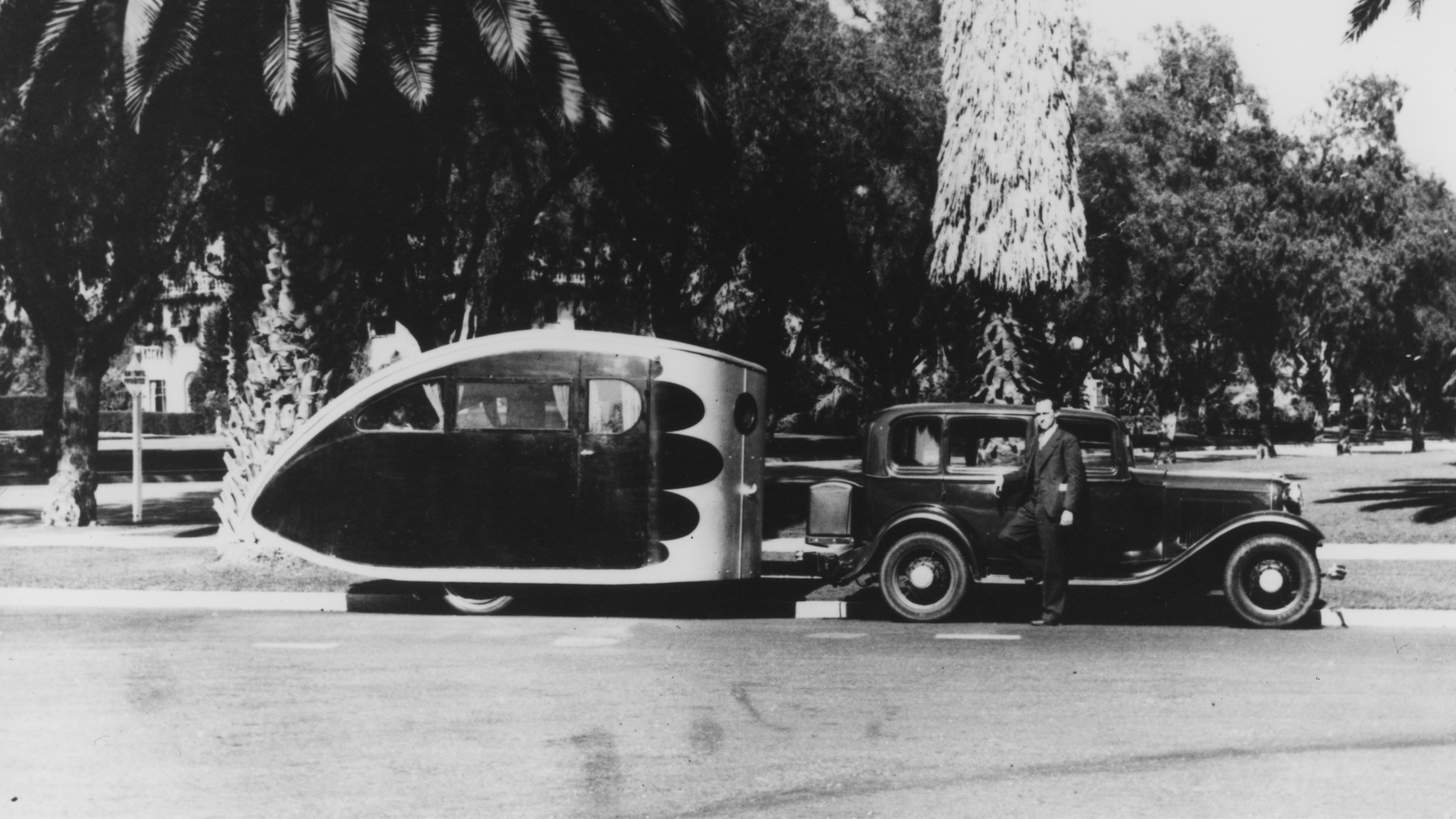 An Airstream Torpedo travel trailer being towed by a vintage car in a tropical area|An old Western Music and Radio Trades Journal|A vintage Airstream Torpedo travel trailer with people sitting outside of the trailer and car.|Two women sitting in lawn chairs next to an Airstream Torpedo travel trailer|An old clip from a newspaper about the Airstream Torpedo travel trailer|Dr. and Mrs. Holman holding up the Airstream Torpedo travel trailer blueprint while standing in front of the Airstream trailer|An Airstream Torpedo travel trailer being towed by a vintage car in a tropical area