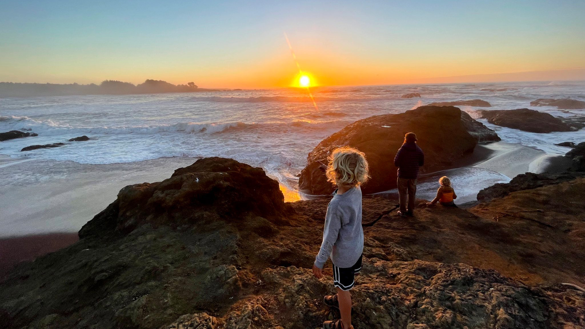Kids and their parent standing on rocks by a body of water during a sunset.|Three campers sitting at a table in their Airstream travel trailer making pasta.|Four kids and their grandma playing Bingo and games in their Airstream travel trailer while they camp.|Four sibling campers sleeping in an Airstream travel trailer in the same bed.|A boy camper grilling in the evening while his family camps in their Airstream travel trailer.|Four kids sitting around a campfire while they camp in their Airstream travel trailer.|Three kids playing outside on a mat under the awning of an Airstream travel trailer while another camper gets a bike out.|Children sitting on a blanket in the sand coloring.|Two kids playing in the sand with sticks next to a body of water.|An Airstream travel trailer parked in a woods while a family camps.|Children walking on a path through the mountains|A child climbing a tree in the rainforest.|A child walking on a path through redwood trees.|Kids hiking in the desert.|A boy walking on a path in between rocks that are in the water.|Kids and their parent standing on rocks by a body of water during a sunset.