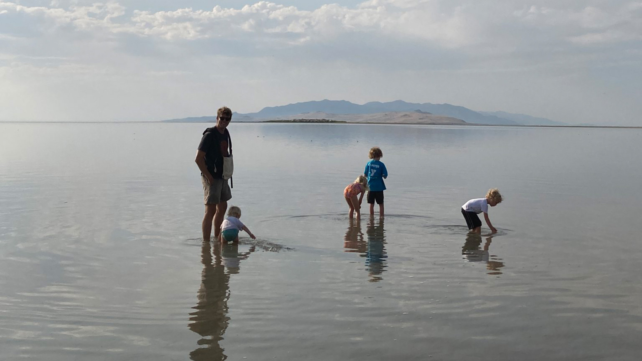 A family walking and playing in shallow water with a fog in the background.|An airstream travel trailer in the mirror of a tow vehicle.|3 kids standing and sitting in a desert looking out at the view on a mountain|2 kids hooking up Airstream travel trailer with their dad in the desert.|3 kids climbing in a tree in a woods.|Three kids running down a little path in a grassy field in the moutains.|Airstream travel trailer parked next to trees during a sunset that is orange and pink.|Two kids sitting on rocks in the middle of a river that is surrounded by trees and a blue sky.|A little kid walking around arches out west.|A family walking and playing in shallow water with a fog in the background.