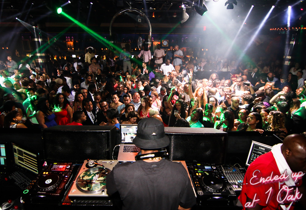 Vibrant nightclub scene at 1 Oak Las Vegas, packed with lively crowd under dynamic green lights, viewed over the shoulder of a DJ at his console.