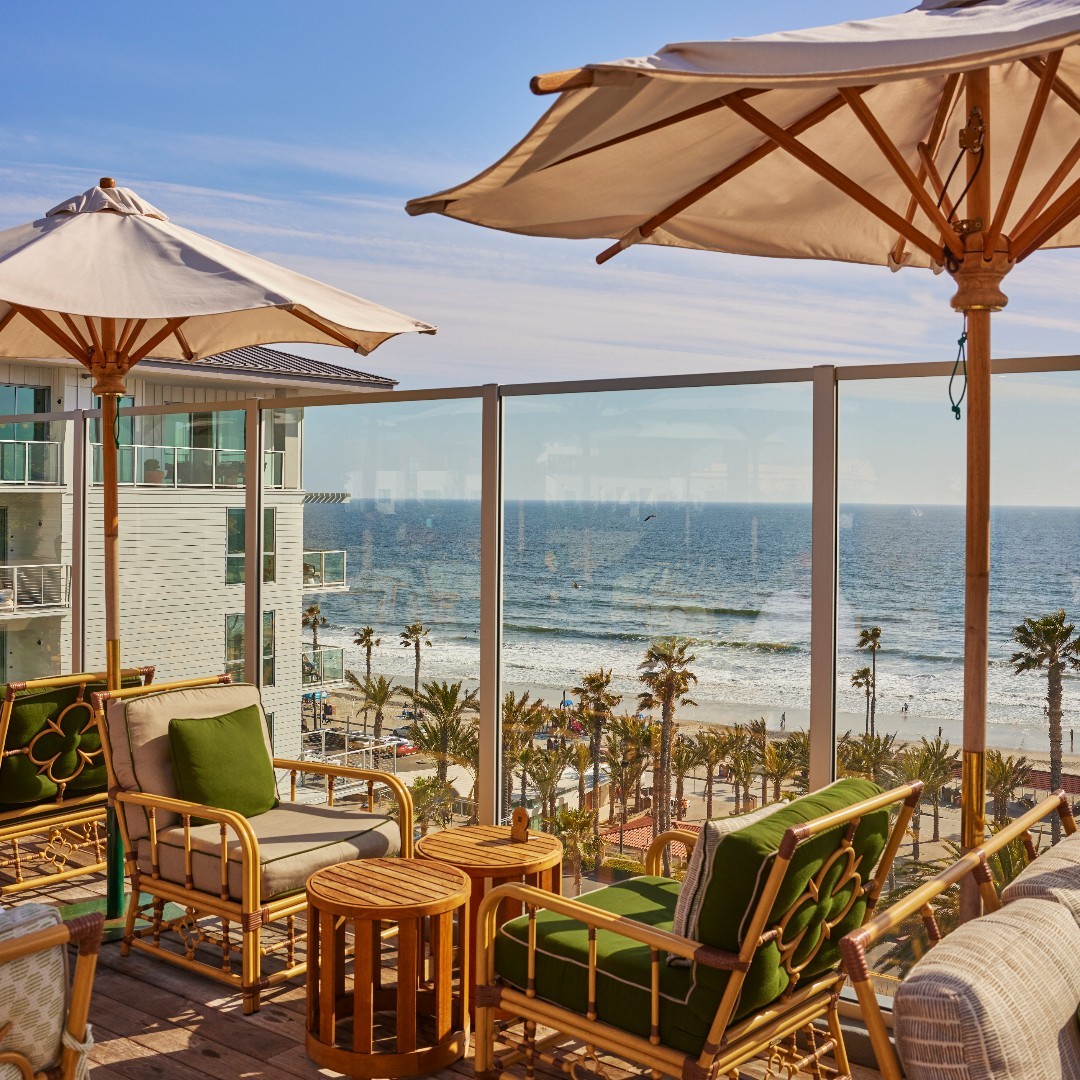 Rooftop lounge at Mission Pacific in San Diego featuring cozy rattan seating with green cushions, elegant white umbrellas, and a stunning view of the sunlit beach and ocean.