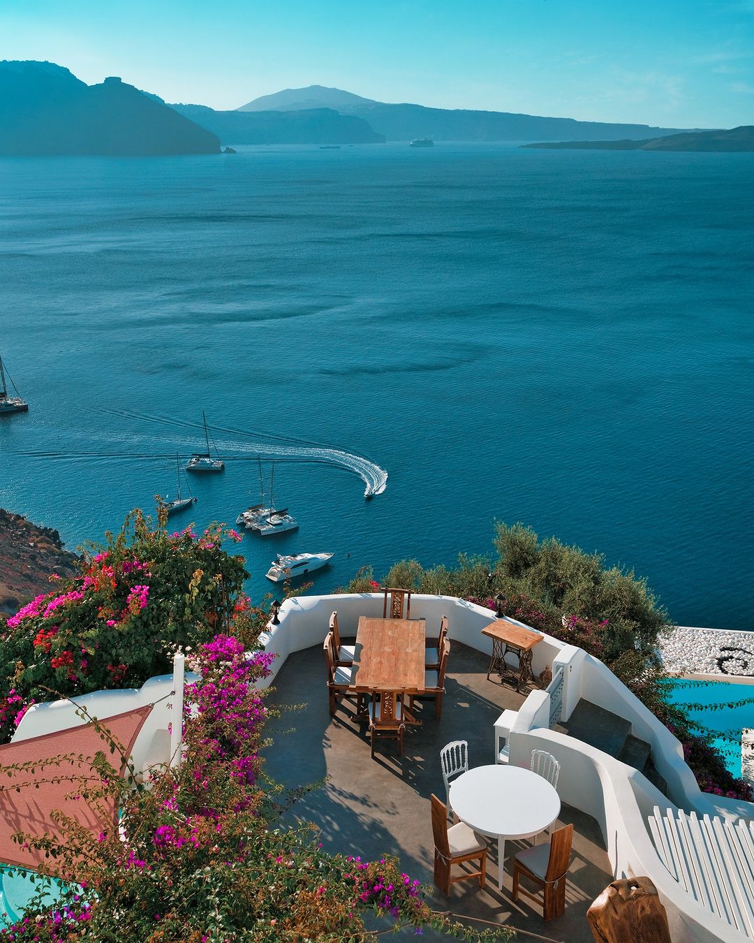 Outdoor restaurant in Santorini with elegant white decor, overlooking a sparkling blue sea and vibrant flowers, ideal for a scenic dining experience.