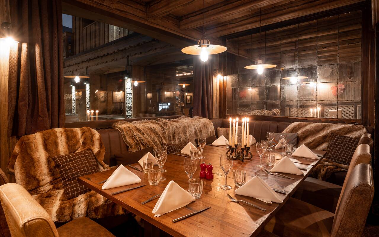 Cozy, warmly-lit interior of Le 80 Meribel restaurant featuring plush seating with fur throws, elegantly set tables with candles, and rustic wooden décor.