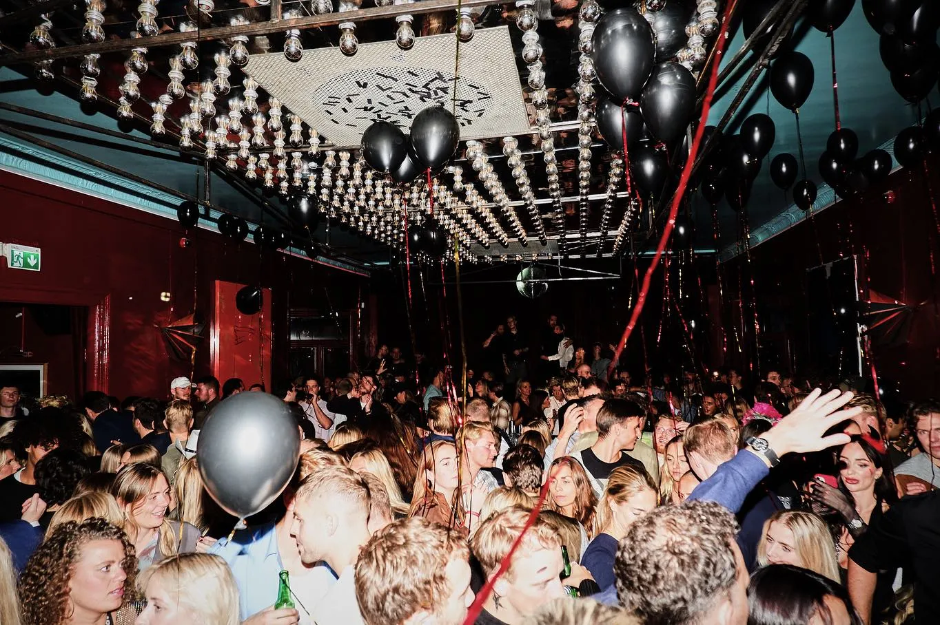 Vibrant nightclub scene at Sø Pavillionen Copenhagen, bustling with a lively crowd under a ceiling adorned with hanging bulbs and black balloons.