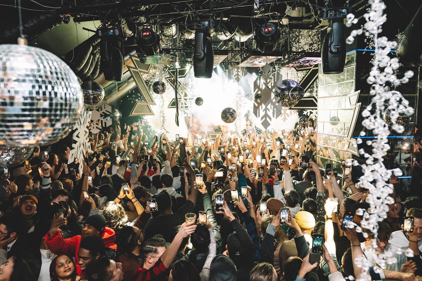 Vibrant nightclub scene at Zeus Tokyo, featuring a crowd of excited partygoers with raised hands and phones capturing the moment, surrounded by dazzling lights and disco balls.