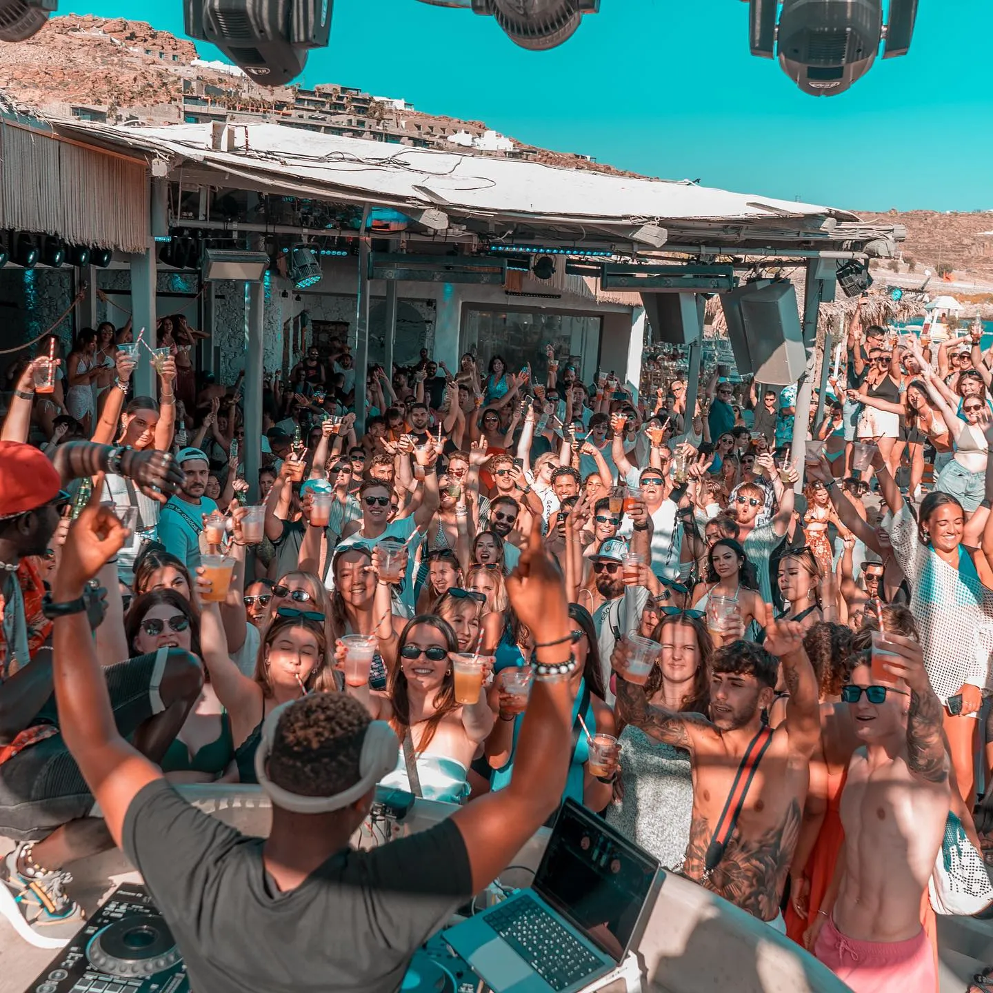 Vibrant beach party at Paradise Beach Club Mykonos with a DJ energizing a cheerful crowd under sunny skies.