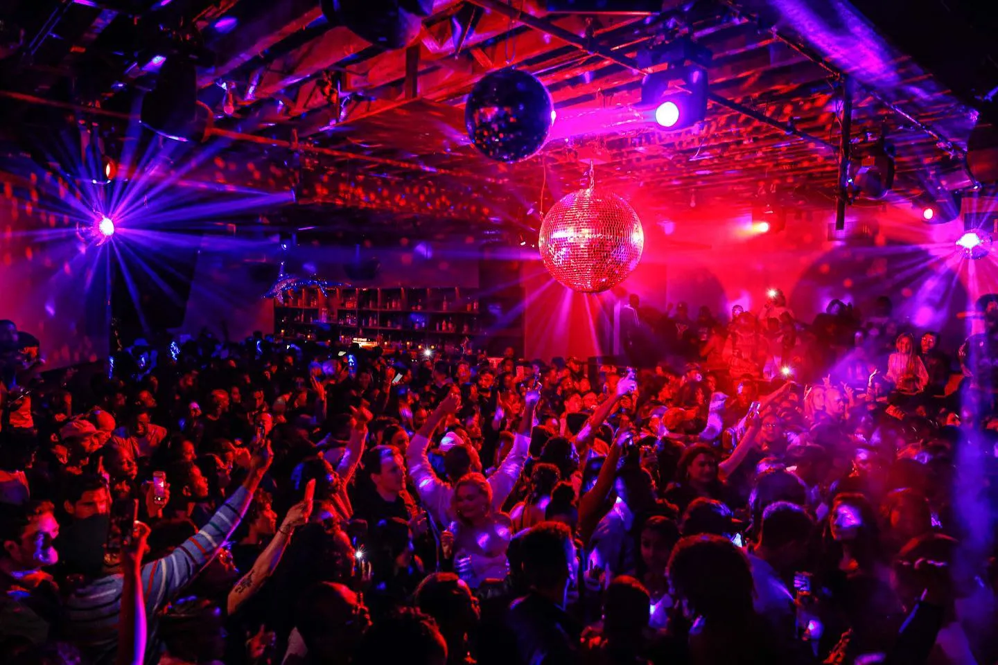 Vibrant nightclub scene with a crowd dancing under pink and blue lights and disco balls at Schimanski New York.