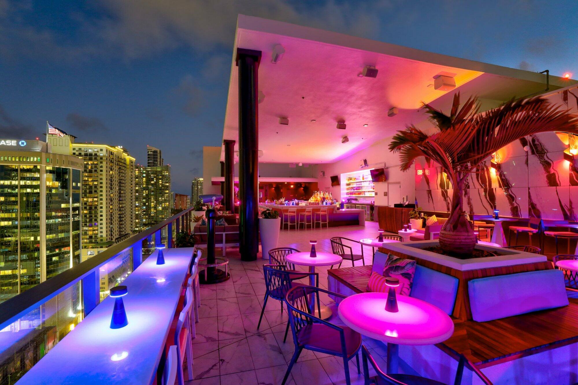 Vibrant Rosa Sky Miami rooftop restaurant at twilight, featuring illuminated neon-lit bar and tables, and a panoramic city view.