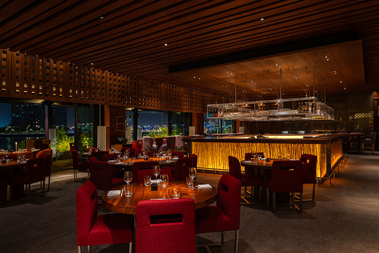 Elegant interior of Zuma Doha restaurant featuring warm lighting, stylish red chairs, and a sophisticated bar, set against a backdrop of large windows showcasing a nighttime city view.