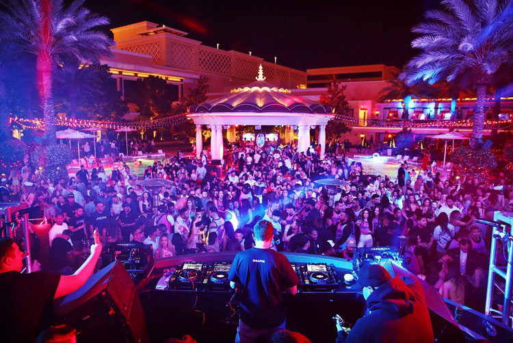 A vibrant scene at XS Las Vegas nightclub, featuring a large crowd enjoying a night under colorful lights with a DJ in the foreground.