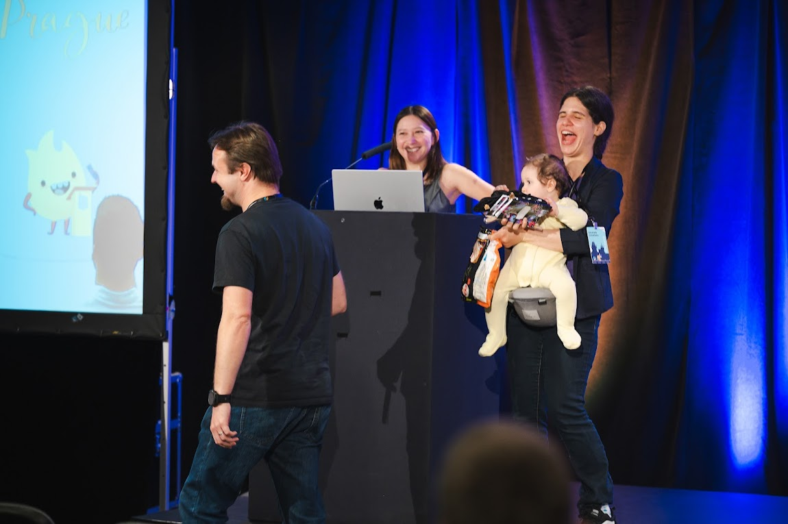 Two women and a baby on stage giving away swag and one man standing in front of them. Everyone is laughing.