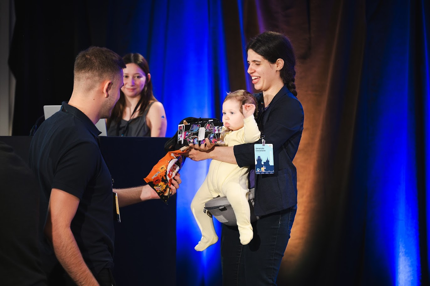 Baby giving away swag to an attendee. Two women speakers on stage. One woman is holding the baby.