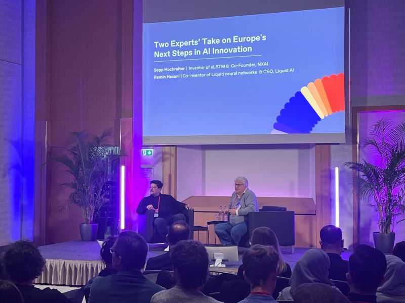 null Two conference speakers sitting on stage, participating in a dialogue while attendees observe. Left in picture Ramin Hasani, Founder of Liquid AI, next to him on stage (right in picture) is Sepp Hochreiter, Founder of NXAI.