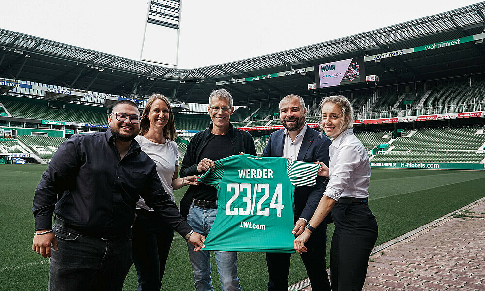 Vetreter:innen von Werder und LWLcom halten ein Trikot im Innenraum des wohninvest WESERSTADION.