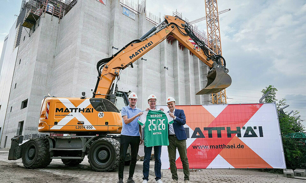 null Ducksch,Filbry und Matthäi-Vertreter vor einem Bagger auf einer Baustelle mit Werder-Trikot in der Hand