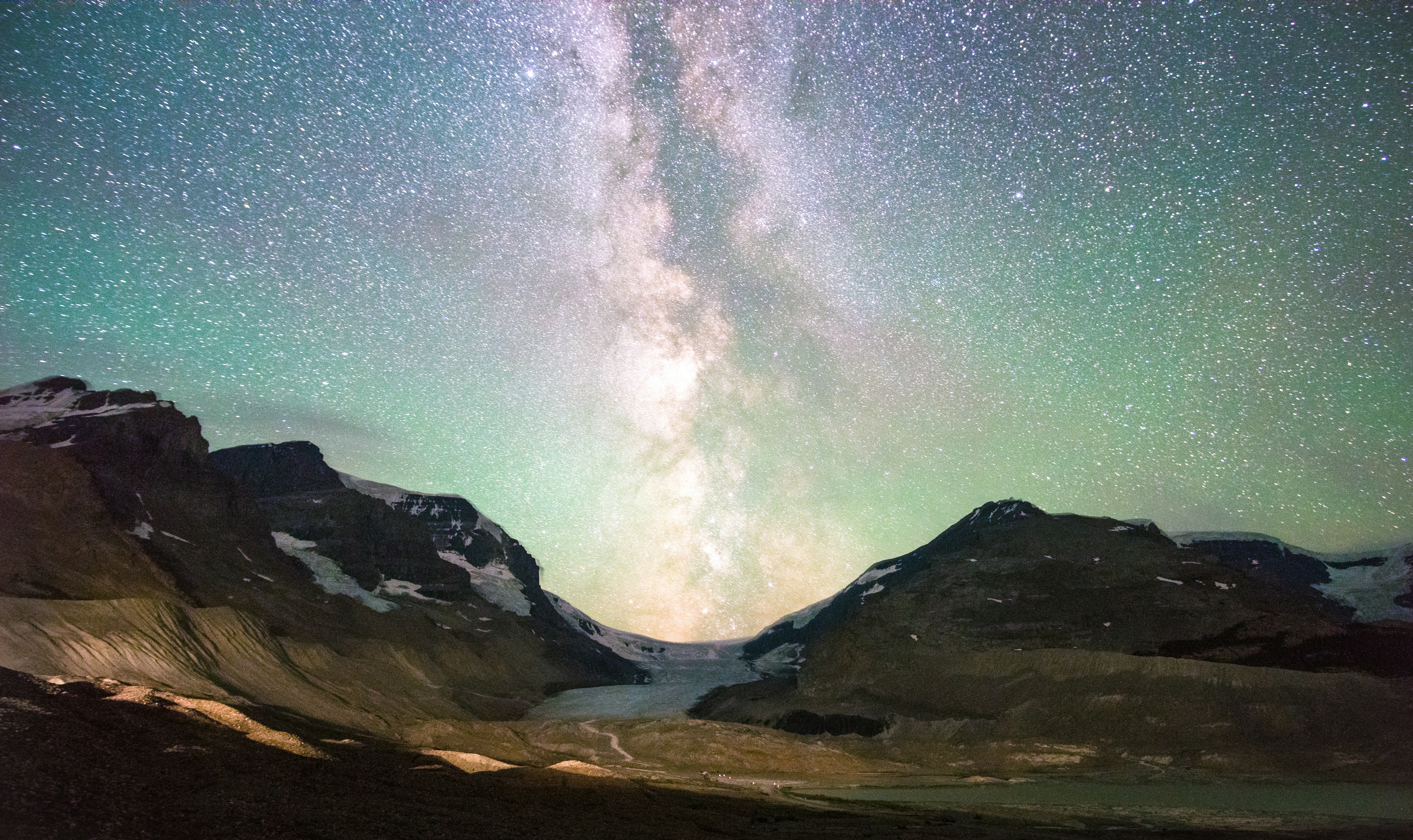 Milky Way core rising above a glacial mountain valley under a green-tinted, star-filled night sky.