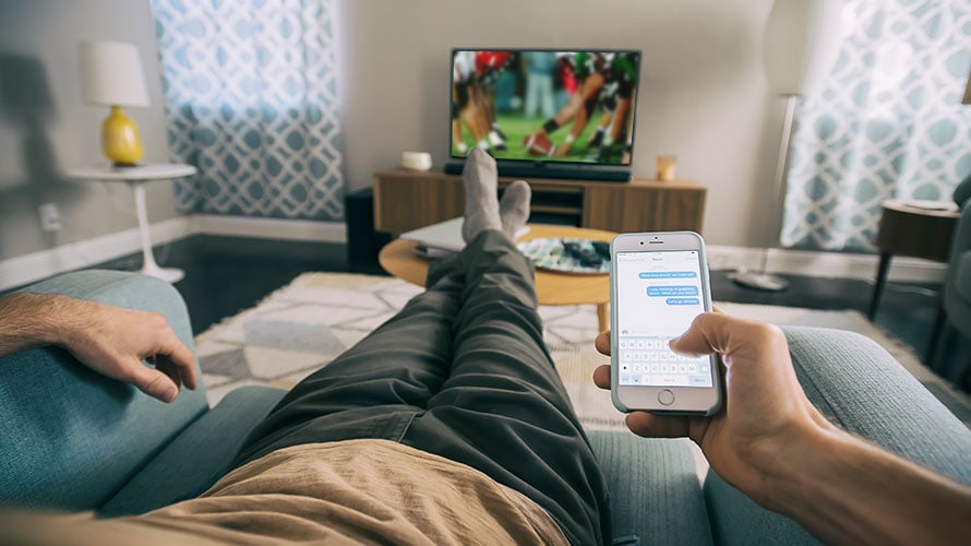 mobile-user-superbowl-content-2019 Point of view image of someone on the couch watching tv; their cellphone is in their right hand and they are texting someone