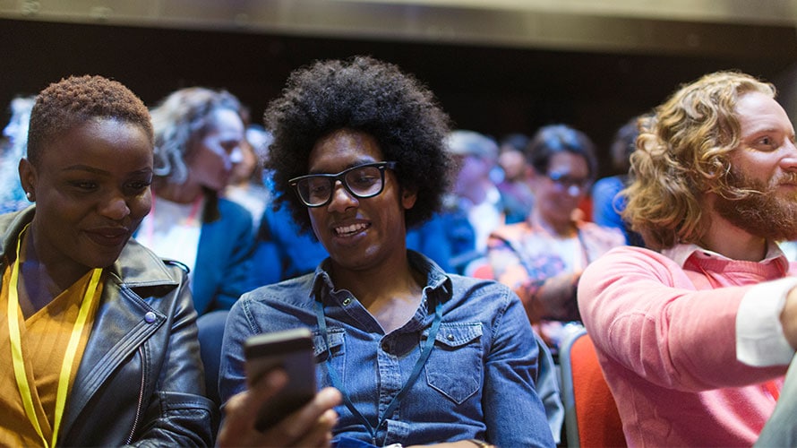 mobile-create-successful-event-content-2019 a person sits in the crowd at an event; the person in the picture is on his phone sharing something next to the attendee next to the person holding the phone