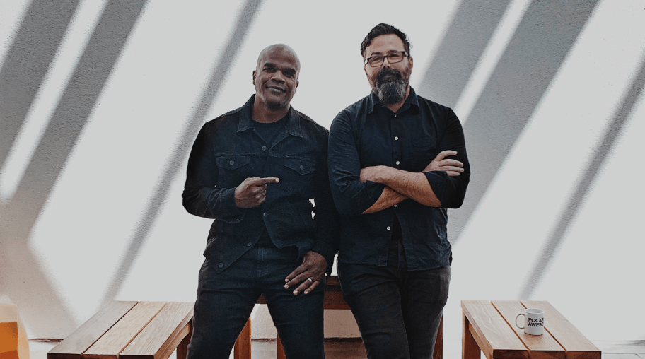 Screen-Shot-2019-05-30-at-1.58.30-PM Geoff Edwards and Chuck Monn pose in dark clothes for a photo against a white wall and wooden tables.