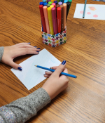 a kid coloring with a pencil and a container of markers nearby