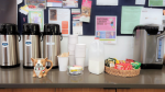carafes of coffee from Blue Canoe on a counter with flyers on a bulletin board behind it