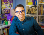 author Jarrett J Krosoczka sits in front of a wall of books he has written.
