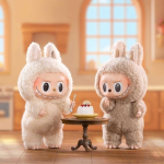 Two labubu toys in a dollhouse room standing next to a table with cake