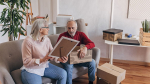 an older couple sitting on a couch going through boxes, the woman showing a photo to a man