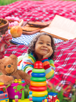 A child with brown curly hair lounges on a red plaid picnic blanket with toys and books around her.