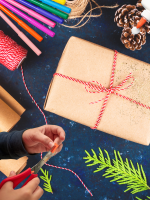 A child's hands are crafting in front of messy and colorful gift-wrapping supplies.