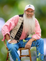 A man with a long white beard in a carnation pink shirt sits on a wooden chair in a field of flowers