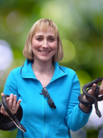 A smiling woman with a blonde bob haircut and a bright blue shirt holds two snakes in front of a mut