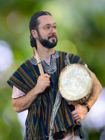 A young man with a beard and horn-rimmed glasses wearing a tunic holds a drum.