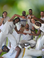 A group of smiling, diverse dancers dressed all in white poses in front of a muted green background.