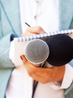 Person in a light blue blazer holding a notepad and two microphones.
