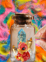 A cute and tiny red dragon toy sits inside of a glass jar with glitter and colorful swirled sand in 