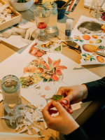 A table with various crafting supplies on it and a person's hands cutting paper above the table. 