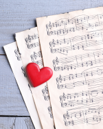 A bright red heart rests on sheet music that is fanned out on a light blue wooden surface.